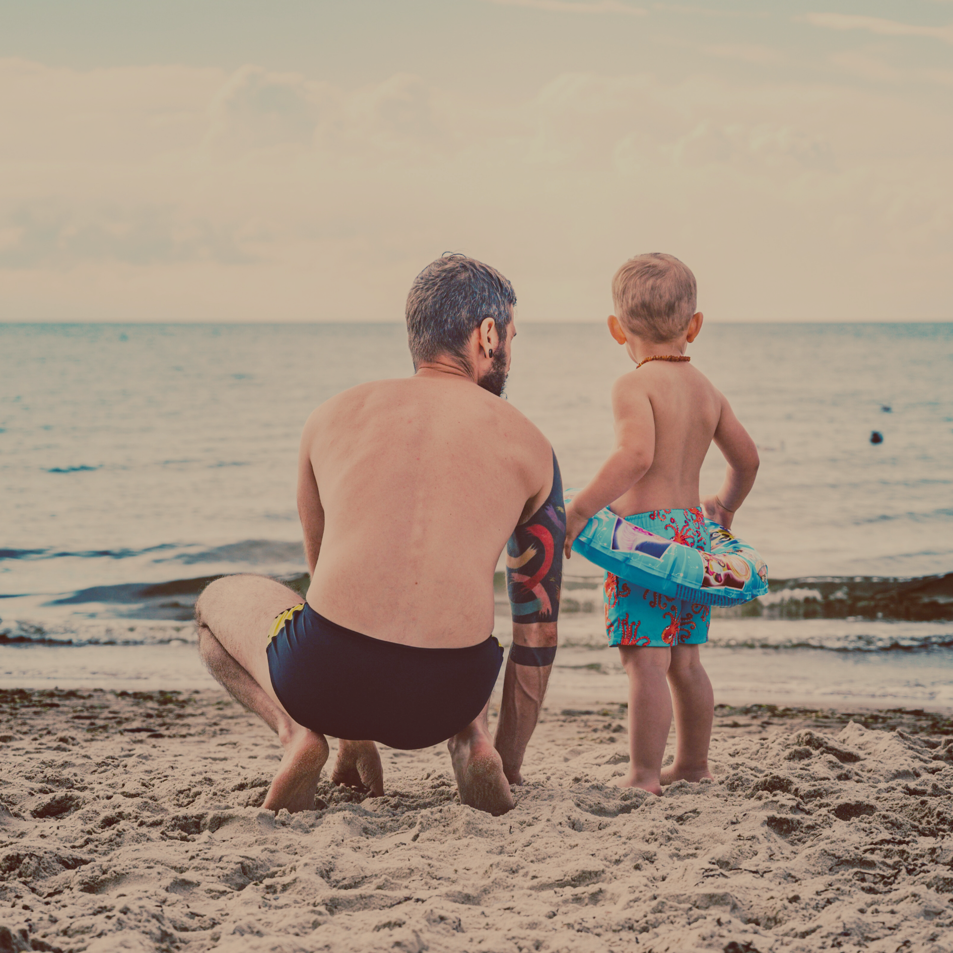 Father crouched down talking to his son as they both look out to the water at the beach.